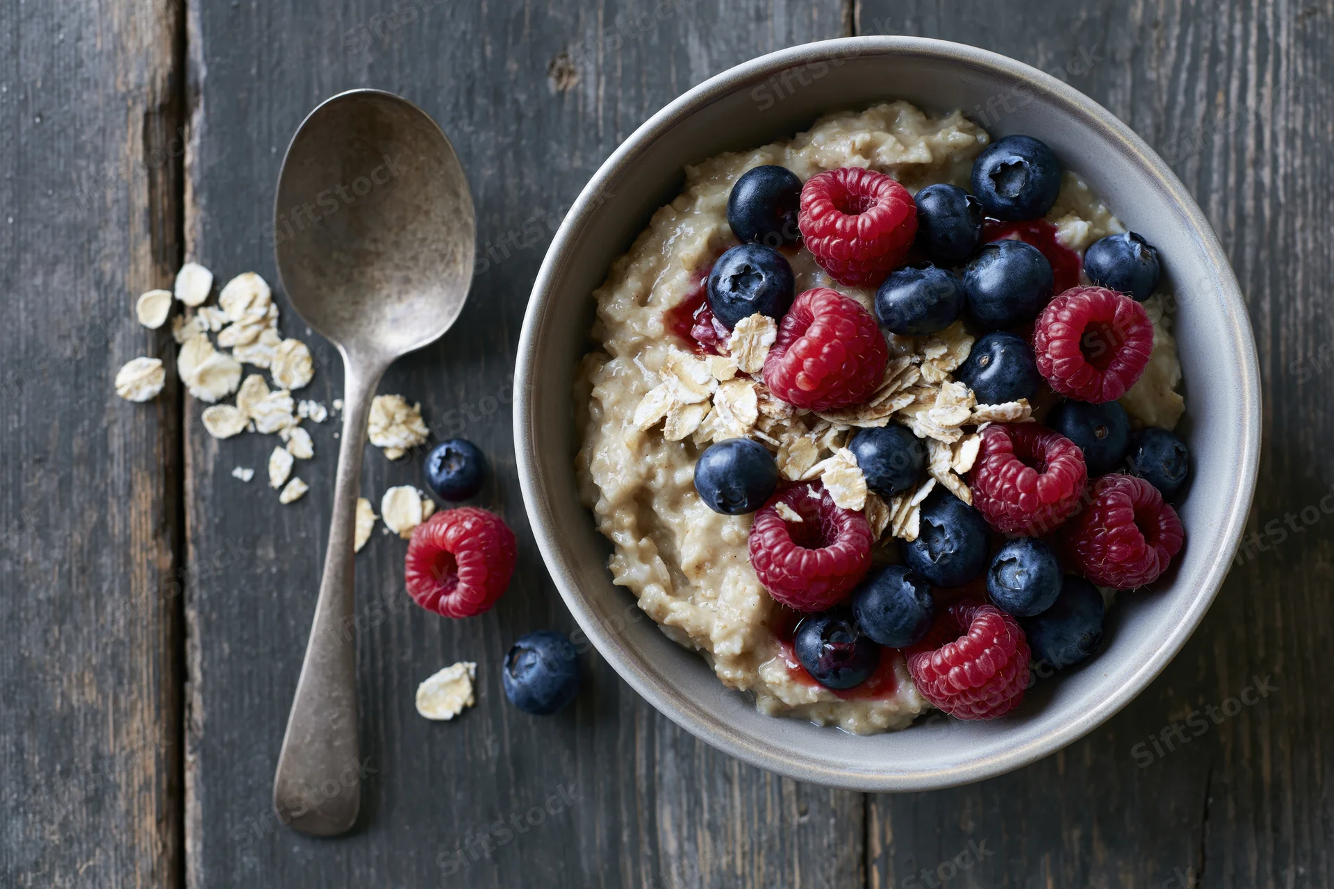 Ceramic bowl of oatmeal porridge topped with fresh raspberries and blueberries on dark wooden table with spoon and scattered oats