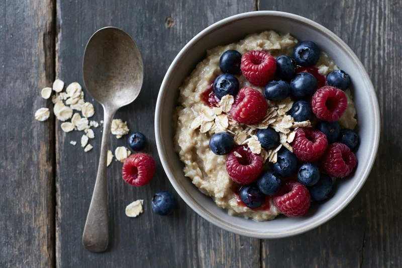 Ceramic bowl of oatmeal porridge topped with fresh raspberries and blueberries on dark wooden table with spoon and scattered oats