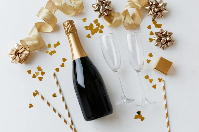 Champagne bottle and two empty flutes with gold ribbons and confetti on white surface