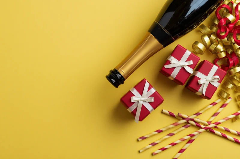 Champagne bottle and red gift boxes with party straws and ribbons on yellow background