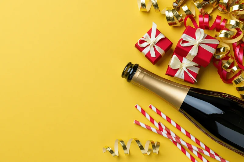 Champagne bottle lying on its side next to red gift boxes and party streamers on yellow background