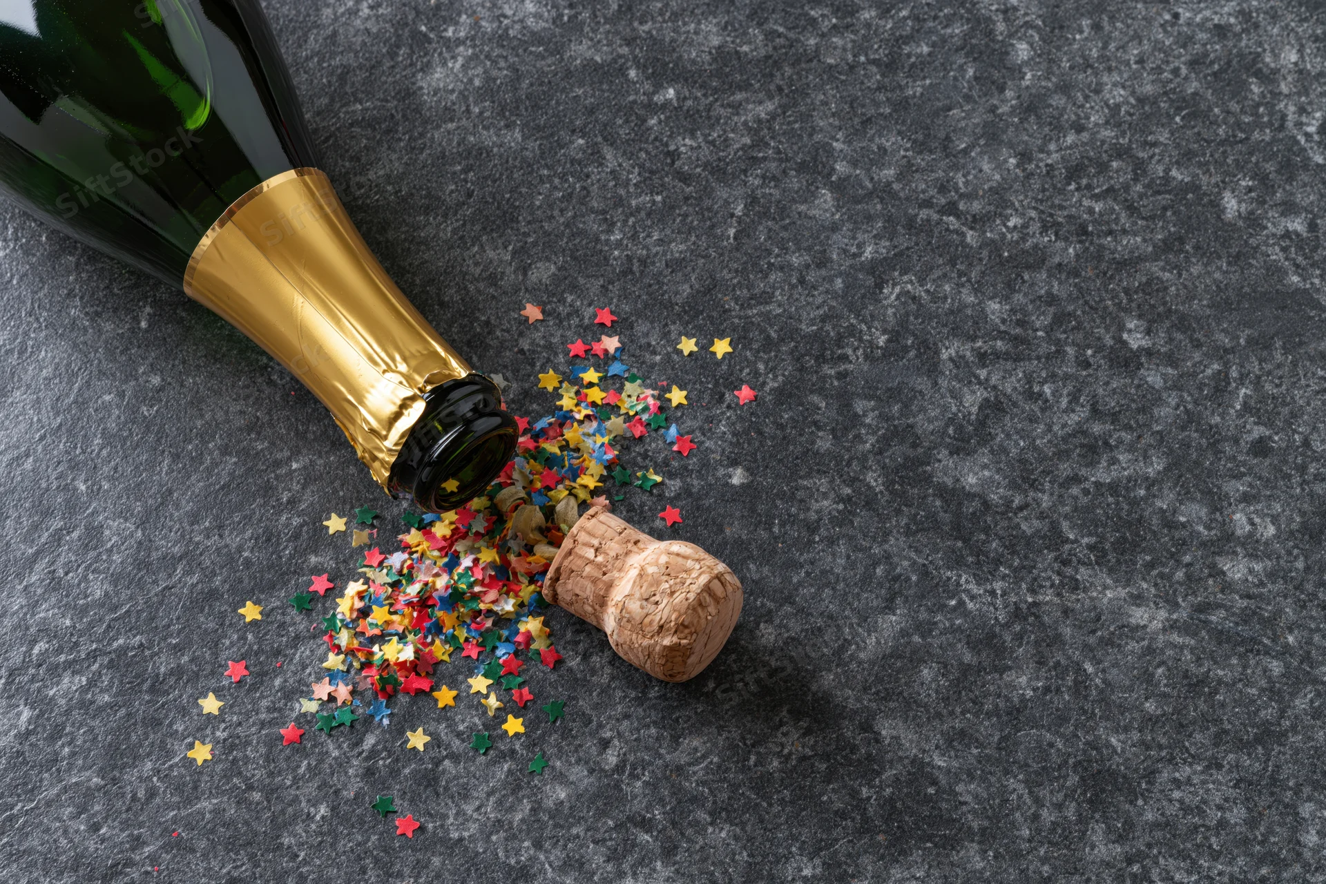 Champagne bottle lying on its side with cork and scattered star-shaped confetti on dark stone surface