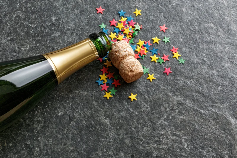 Champagne bottle lying on its side with cork and colorful star confetti on slate surface
