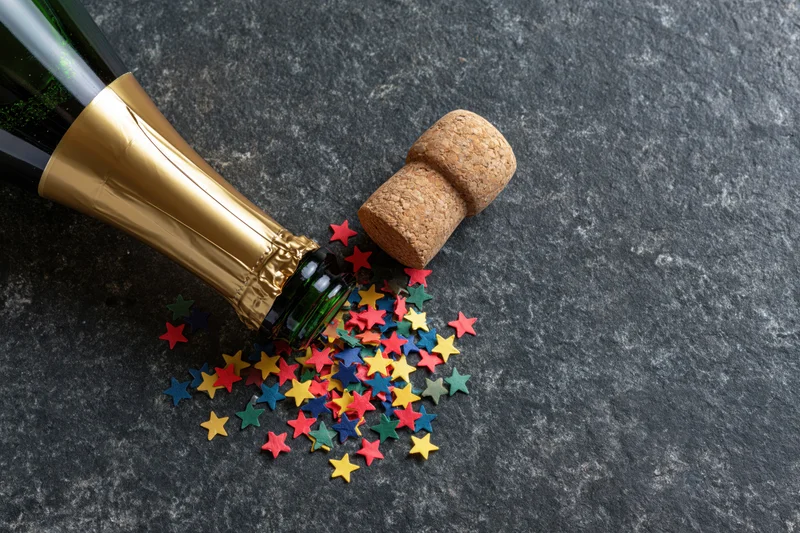 Champagne bottle lying on its side with popped cork and scattered star-shaped confetti on dark textured surface