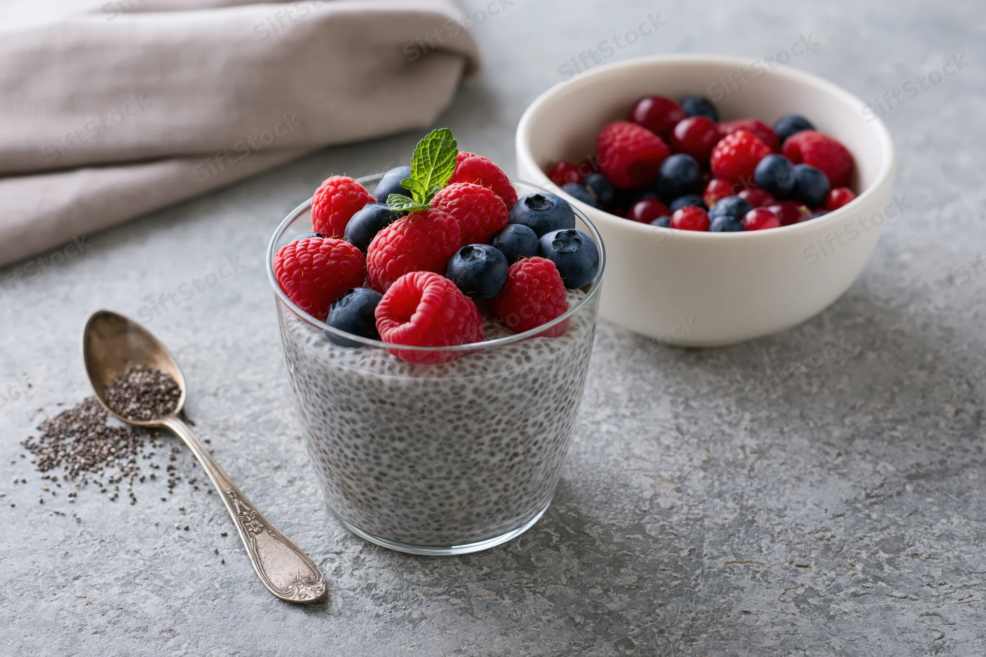 Chia seed pudding in a glass topped with raspberries and blueberries on a gray countertop with a spoon and bowl of berries nearby