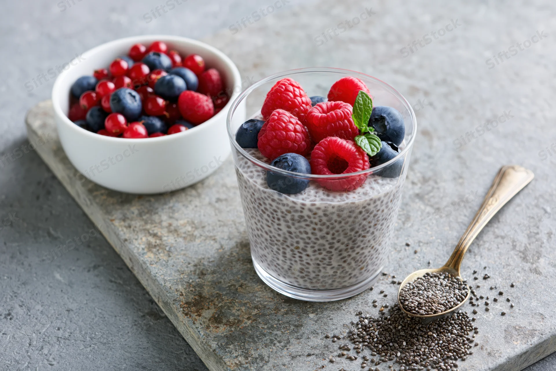Chia seed pudding in a glass topped with raspberries and blueberries on a stone slab with a spoon and bowl of berries nearby