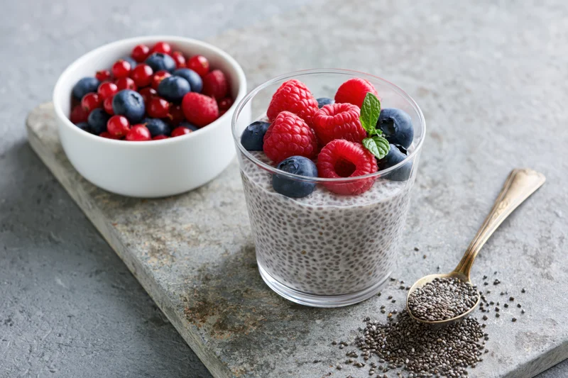 Chia seed pudding in a glass topped with raspberries and blueberries on a stone slab with a spoon and bowl of berries nearby