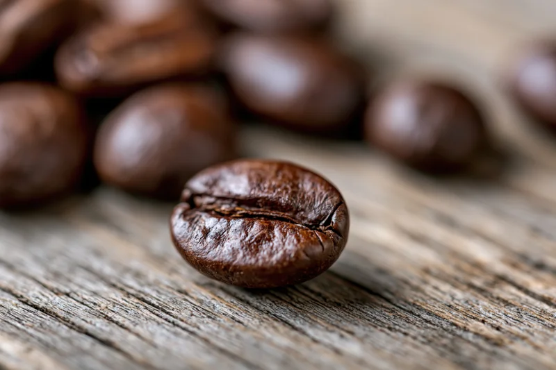 Roasted coffee bean resting on a weathered wooden surface with scattered beans in soft focus background