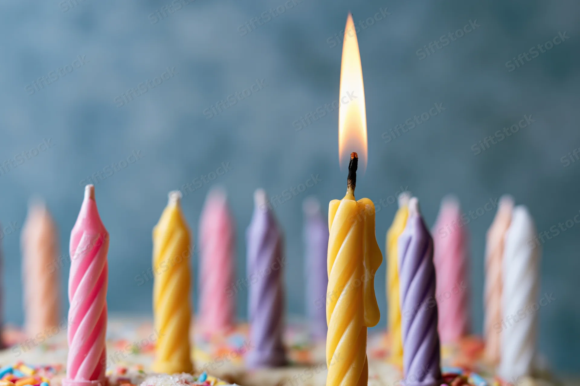 Close-up of colorful twisted birthday candles on a cake with one lit flame against a blurred blue background