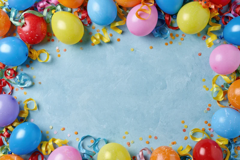 Colorful party balloons and curled ribbon streamers arranged in a border around a light blue surface with confetti, leaving central copyspace