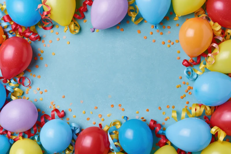 Colorful party balloons and curled ribbon streamers arranged in a frame around a blue surface with confetti