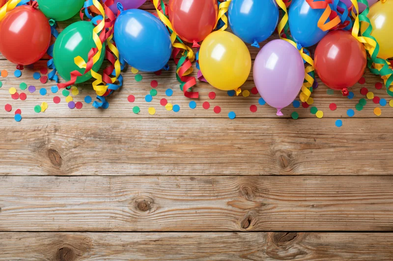 Colorful party balloons and streamers scattered on a wooden table with confetti, top view, copyspace below
