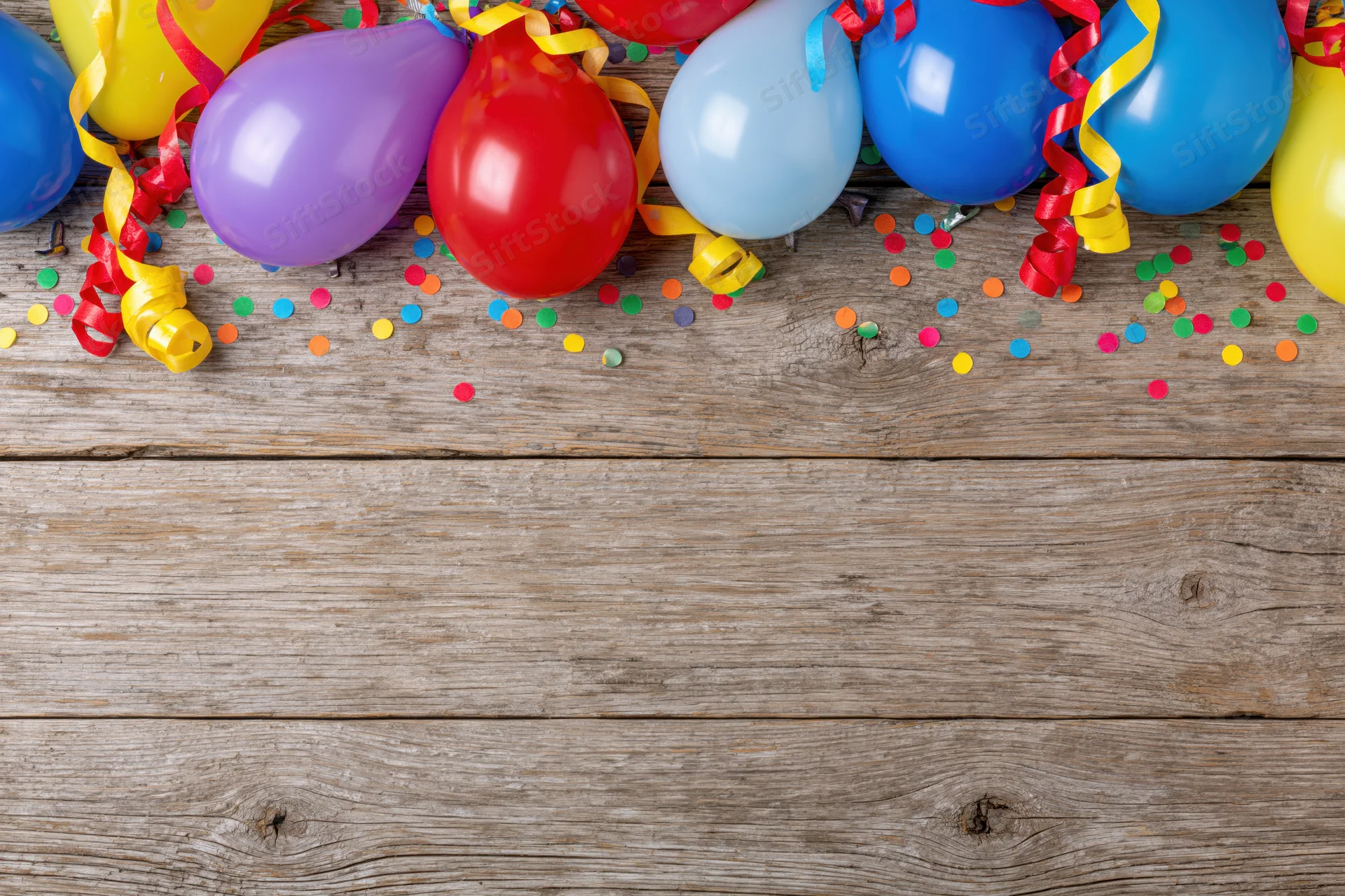 Colorful party balloons and streamers with confetti arranged along the top of a rustic wooden table, leaving copyspace below