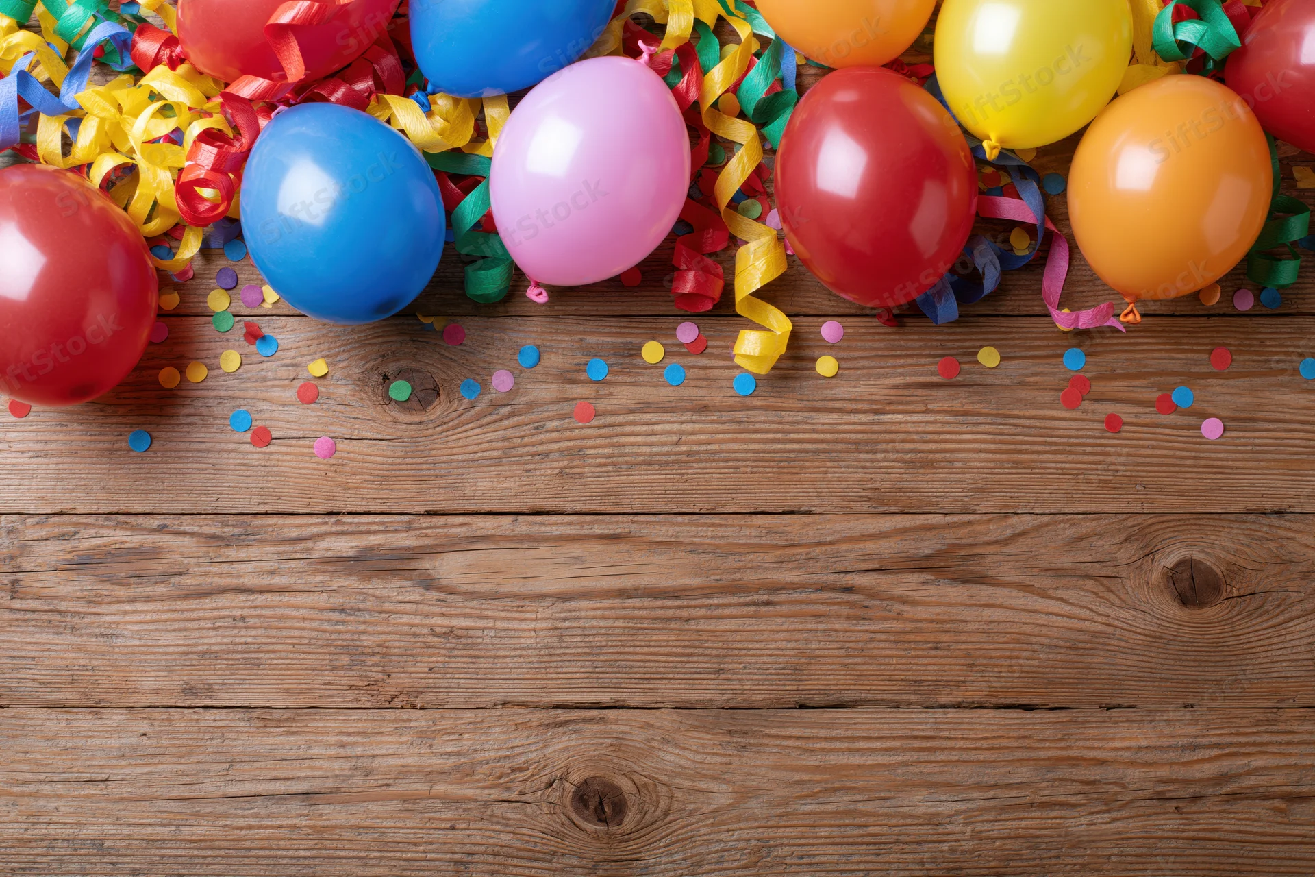 Colorful party balloons and streamers scattered on a wooden table with confetti and copyspace below