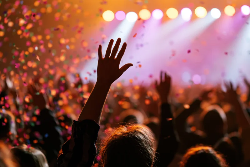 Crowd at concert with arms raised toward stage under colorful lights and falling confetti