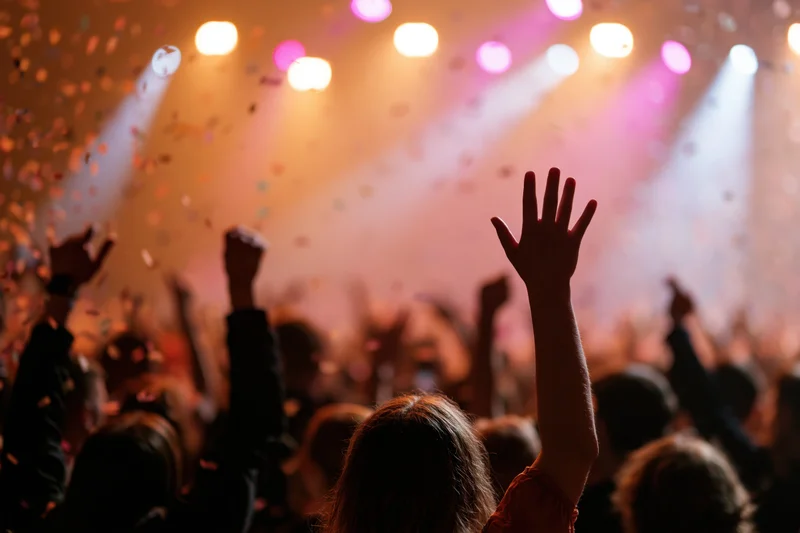 Crowd at concert with arms raised toward stage under colorful lights and falling confetti