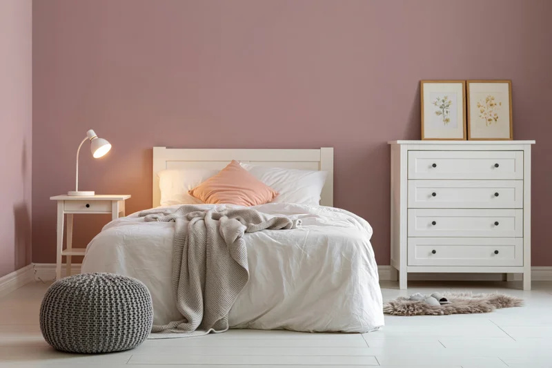 Cozy bedroom with white bed and pink walls featuring nightstand, dresser, and knitted pouf
