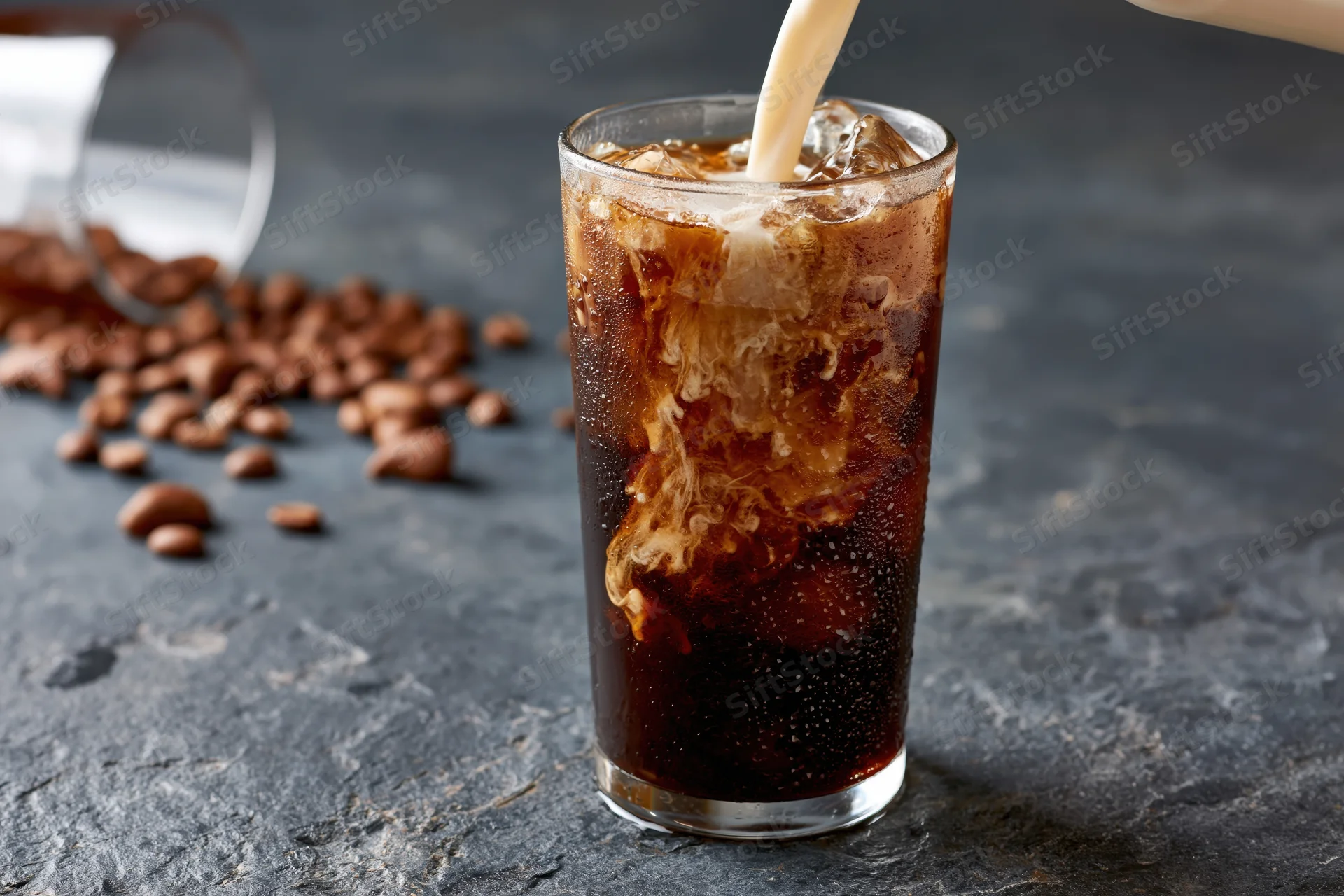 Cream being poured into a tall glass of iced coffee swirling on a dark stone surface with scattered coffee beans nearby