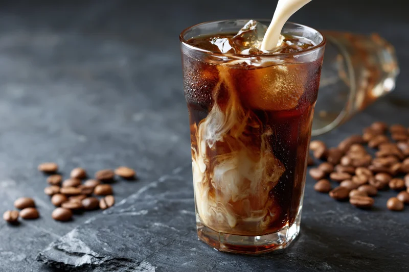 Cream being poured into a tall glass of iced coffee swirling on a dark slate surface with coffee beans scattered around