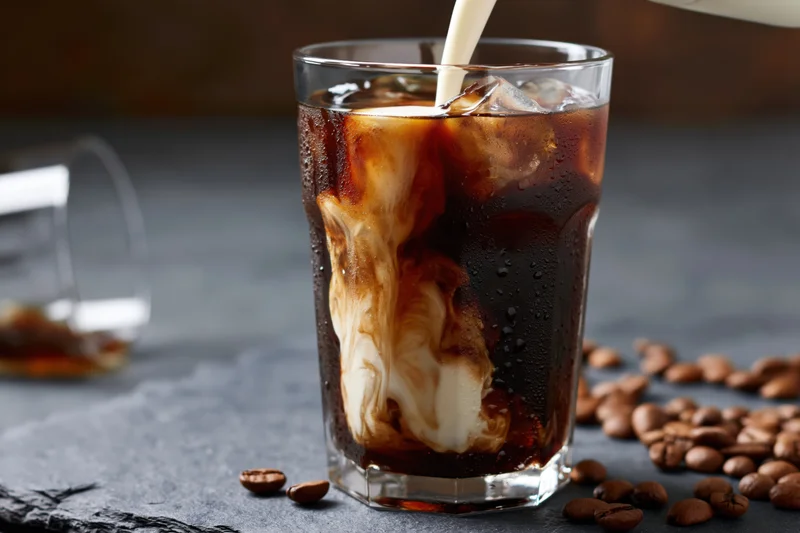 Cream being poured into a tall glass of iced coffee swirling on a slate surface with coffee beans scattered around
