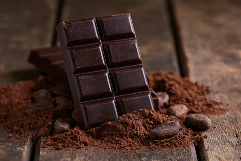 Dark chocolate bar standing upright on wooden surface surrounded by cocoa powder and beans