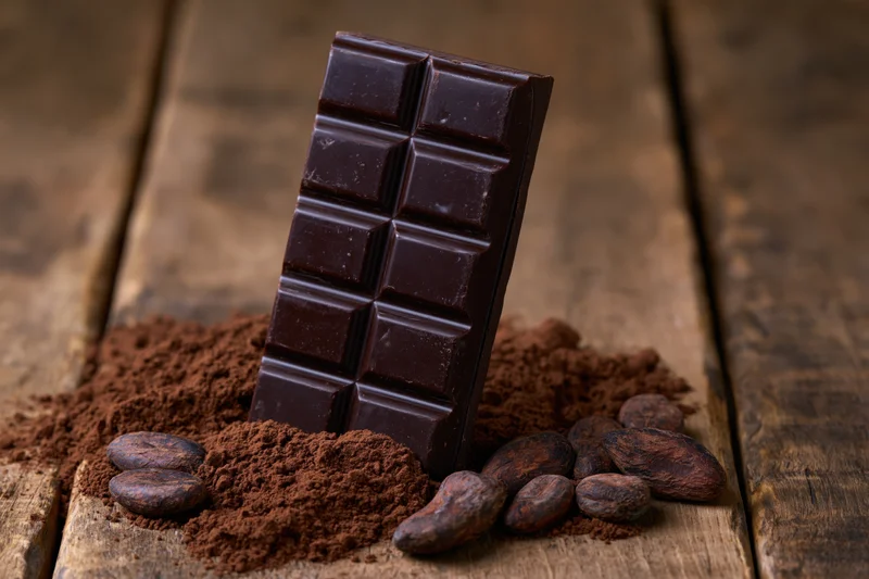 Dark chocolate bar standing upright on wooden surface surrounded by cocoa beans and cocoa powder