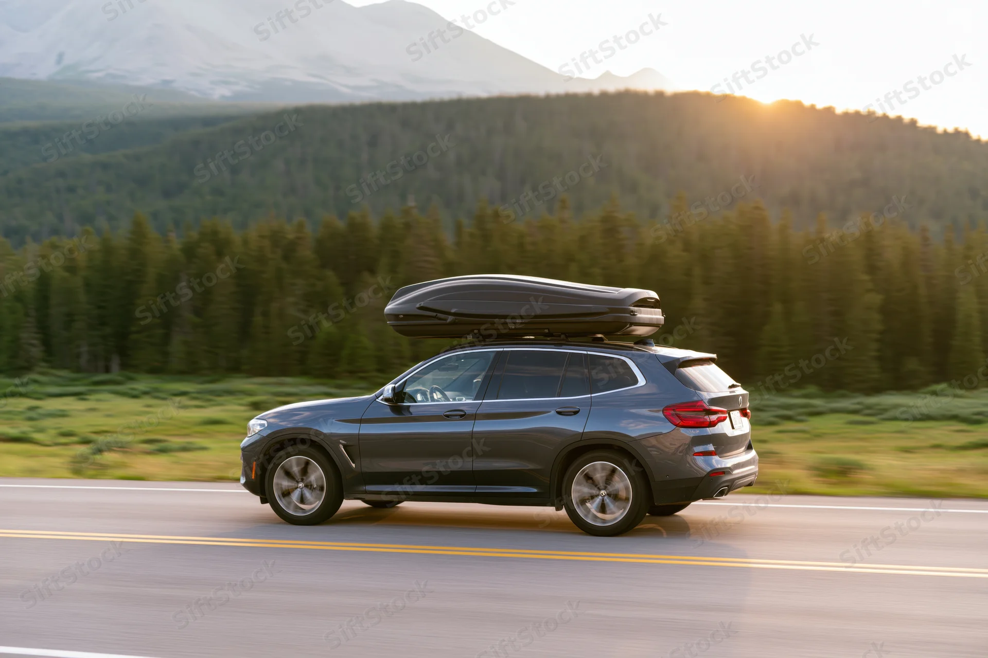 Dark grey luxury suv with black roof cargo box driving on highway past forested mountains at sunset