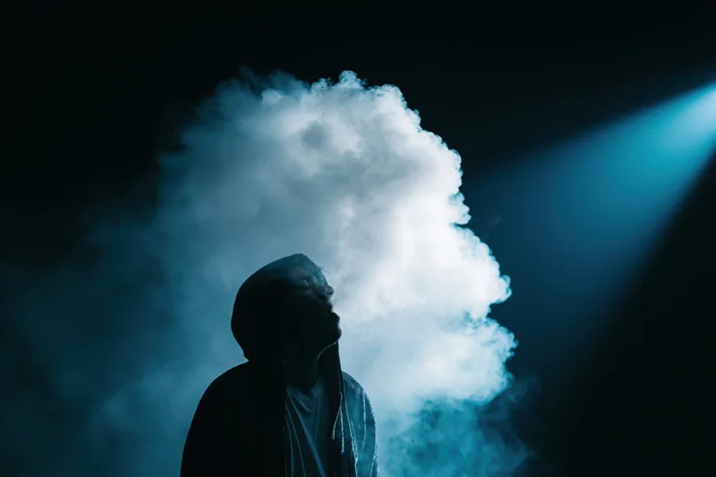 Hooded figure exhaling thick smoke cloud in dark blue-lit space