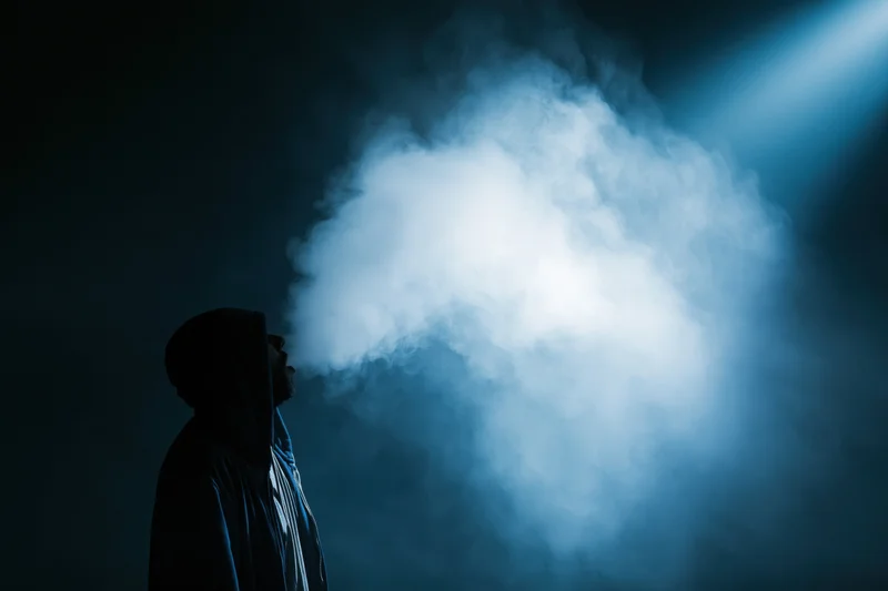 Hooded figure exhaling a large cloud of vapor in a dark blue-lit space