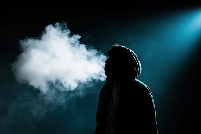 Hooded figure exhaling a large cloud of vapor in a dark blue-lit space