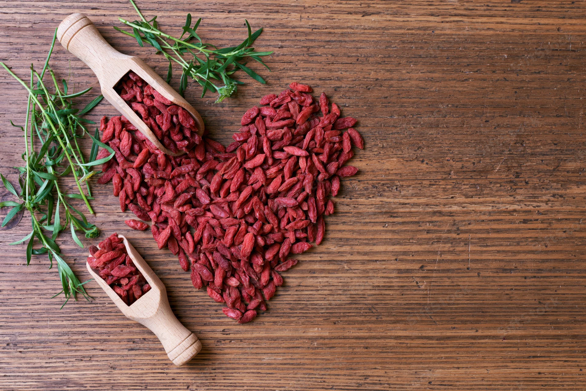Dried goji berries arranged in heart shape on wooden surface with green sprigs and wooden scoops