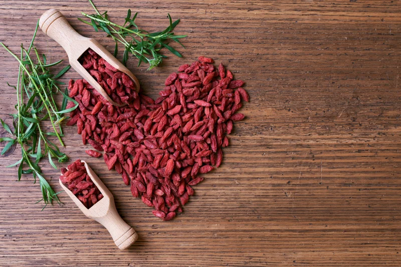 Dried goji berries arranged in heart shape on wooden surface with green sprigs and wooden scoops