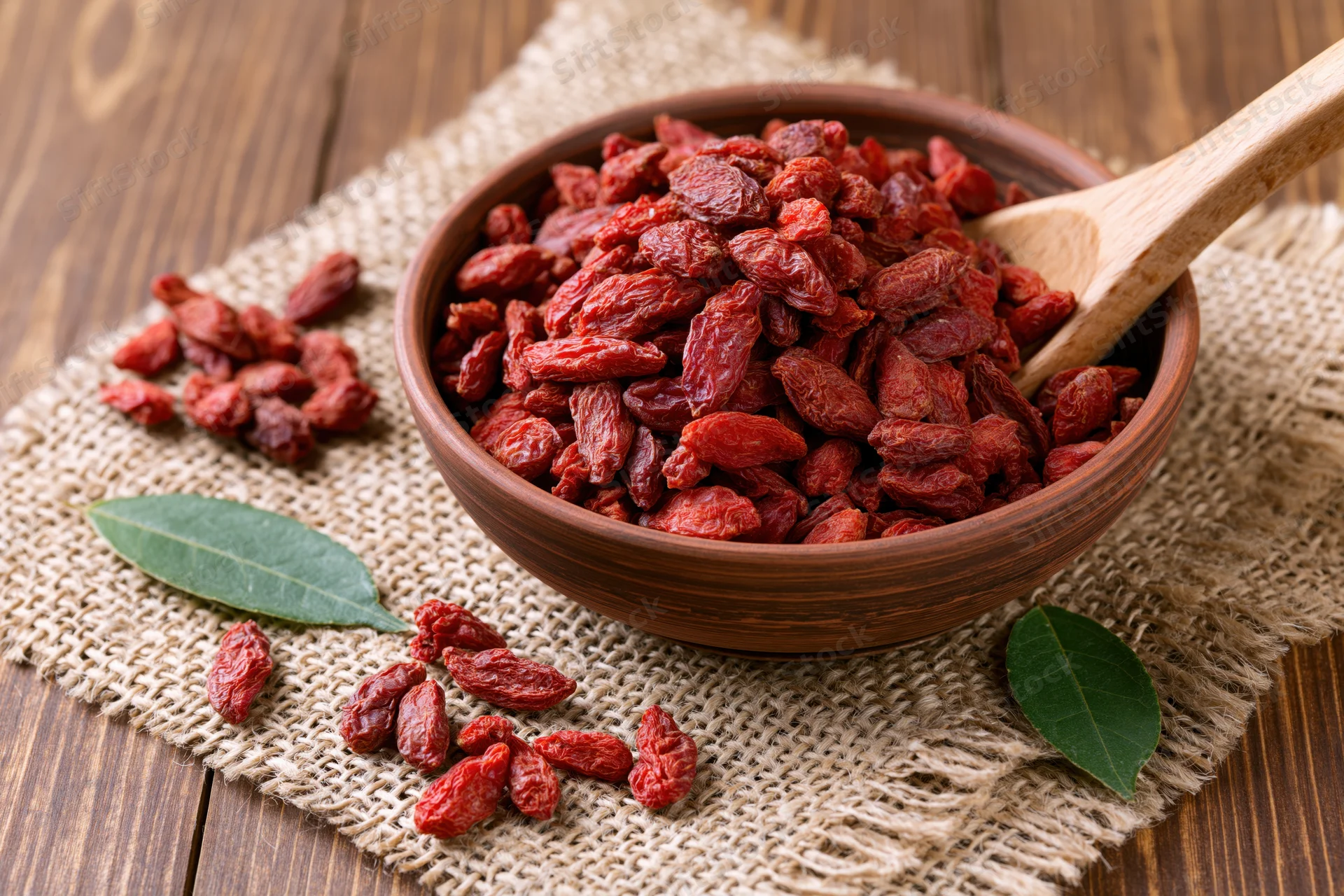 Dried goji berries in a rustic clay bowl with wooden spoon on burlap over wood table