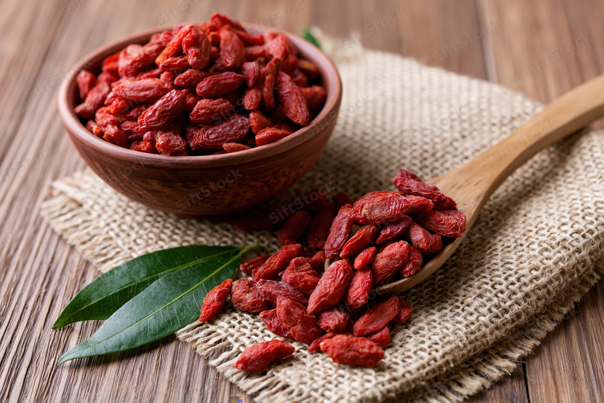 Dried goji berries in a clay bowl and wooden spoon on burlap cloth over wood table