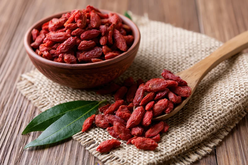 Dried goji berries in a clay bowl and wooden spoon on burlap cloth over wood table