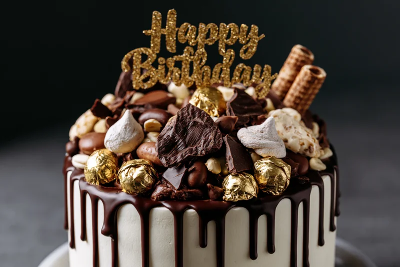 Chocolate drip birthday cake topped with gold foil candies and chocolate pieces on a dark background