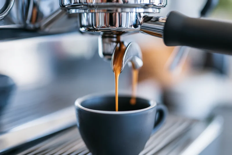 Espresso machine portafilter extracting coffee into a black ceramic cup on a commercial counter