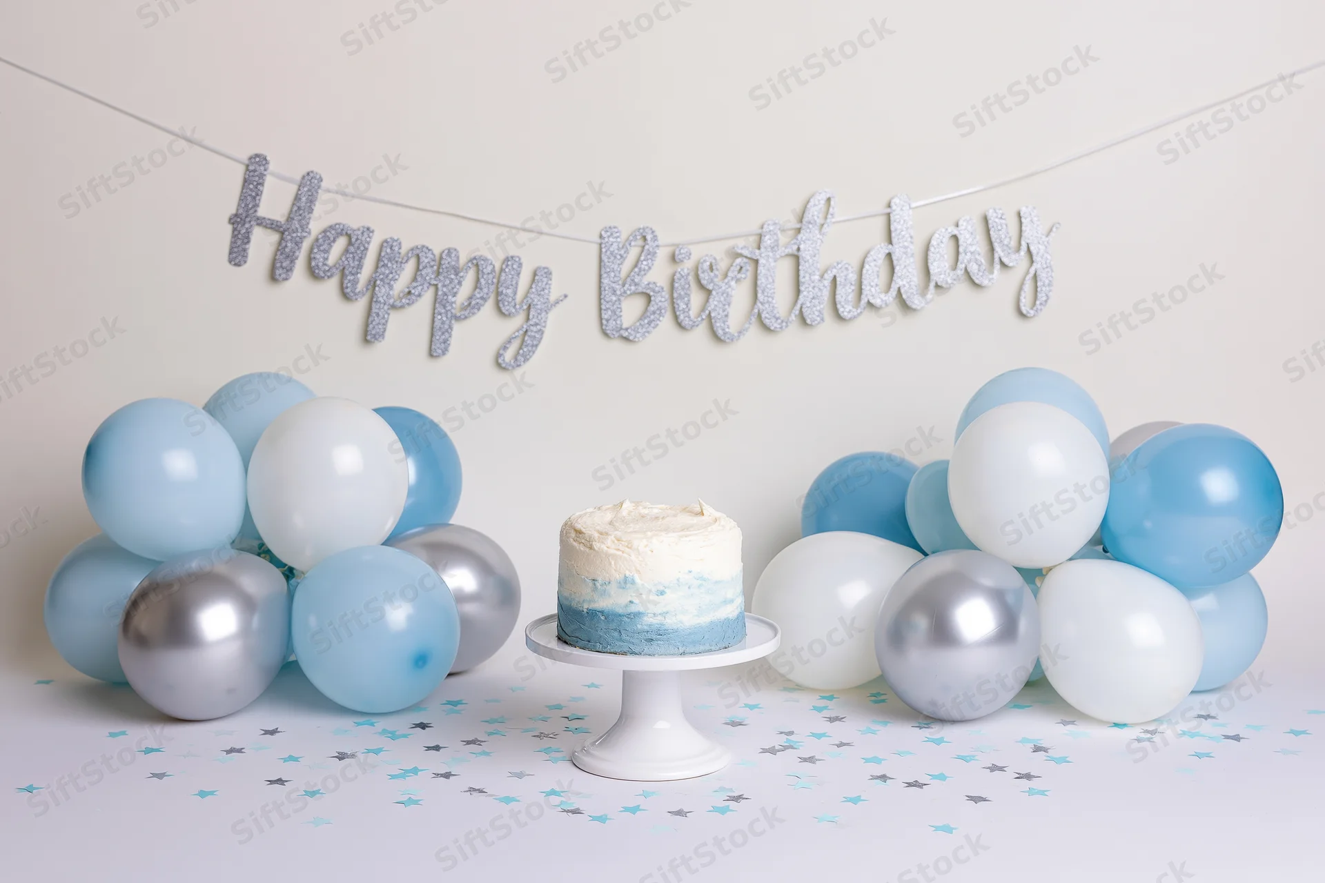 Birthday cake on a stand surrounded by blue and silver balloons with a glitter banner above on a white table with scattered confetti