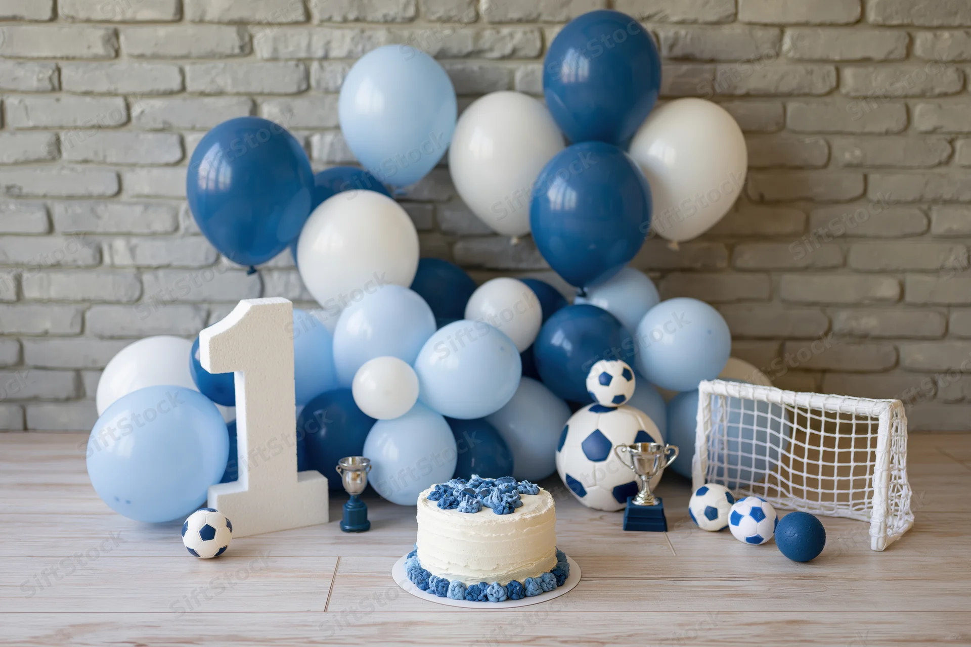 First birthday party setup with blue and white balloons, soccer-themed decorations, cake, trophy, and number one on a wooden table against a brick wall