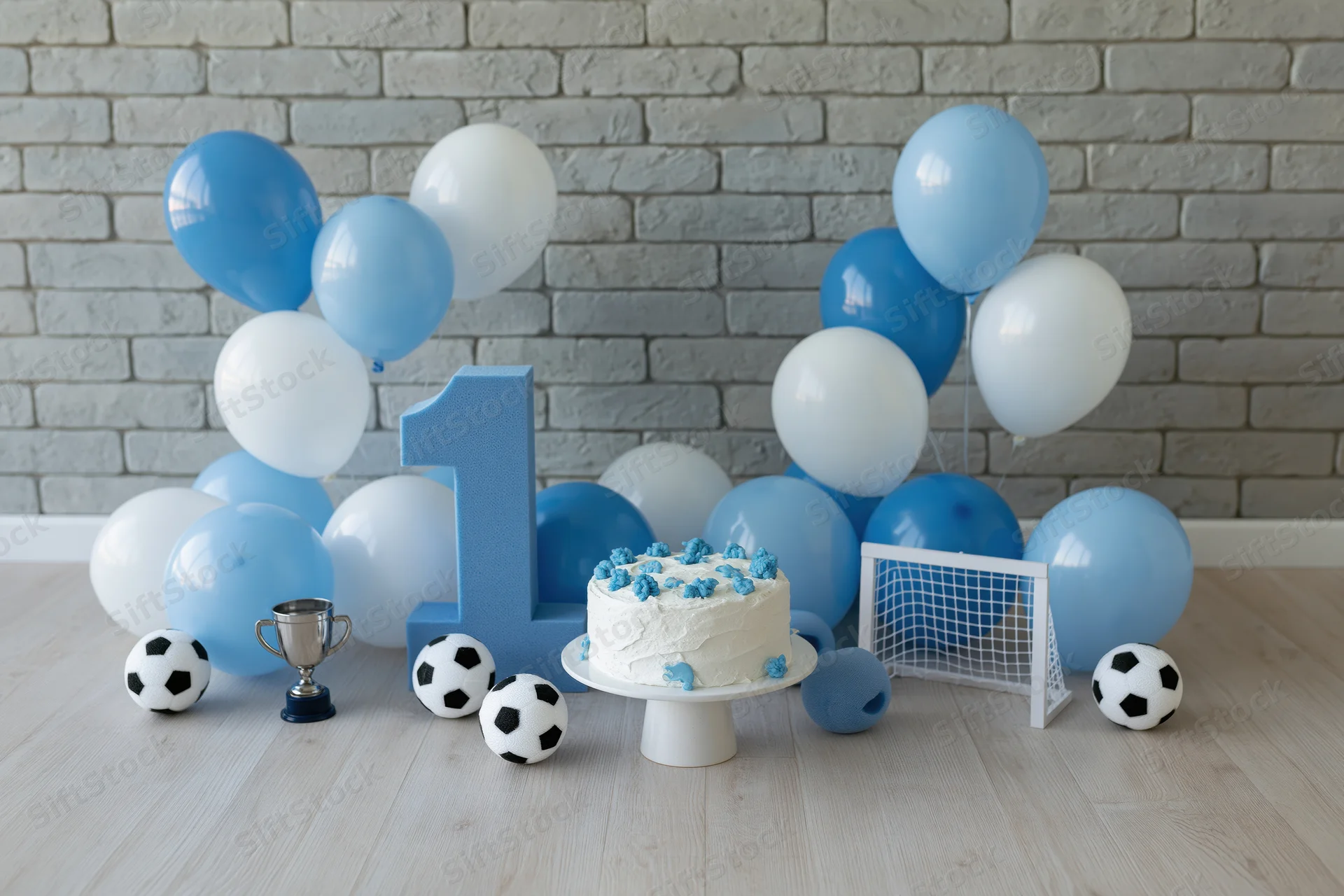 First birthday party setup with cake, balloons, soccer balls and trophy on wooden floor against brick wall