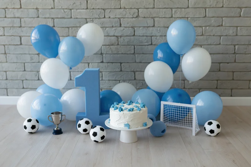 First birthday party setup with cake, balloons, soccer balls and trophy on wooden floor against brick wall