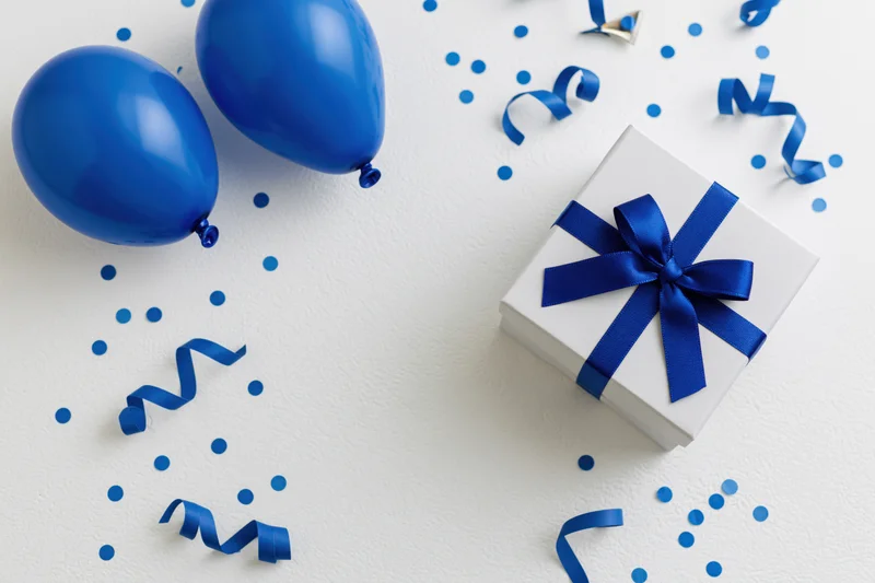 Flat lay birthday arrangement with blue balloons, gift box, ribbon and confetti on white surface