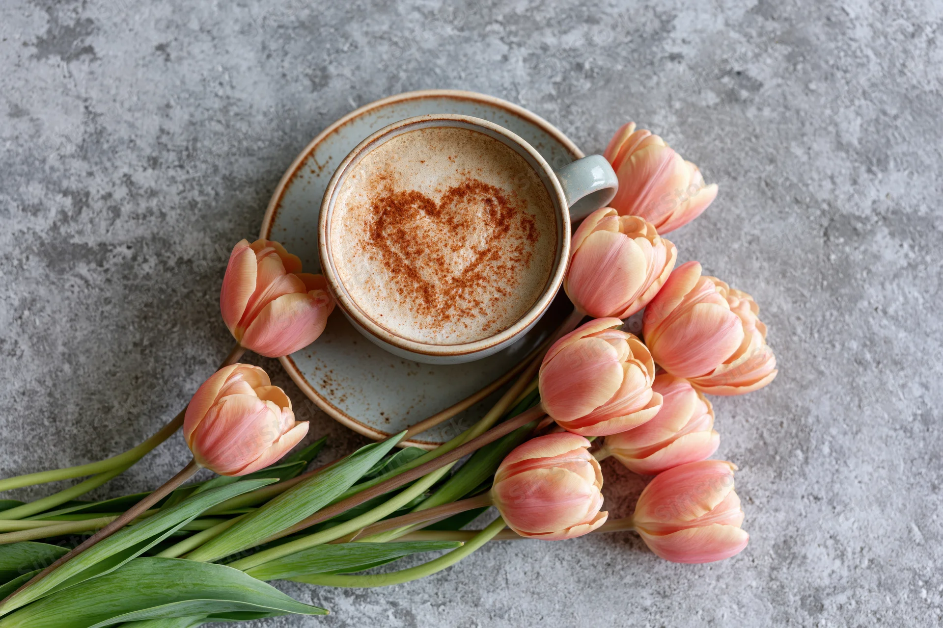 Ceramic cappuccino cup with heart-shaped cinnamon design and pink tulips arranged on gray concrete surface