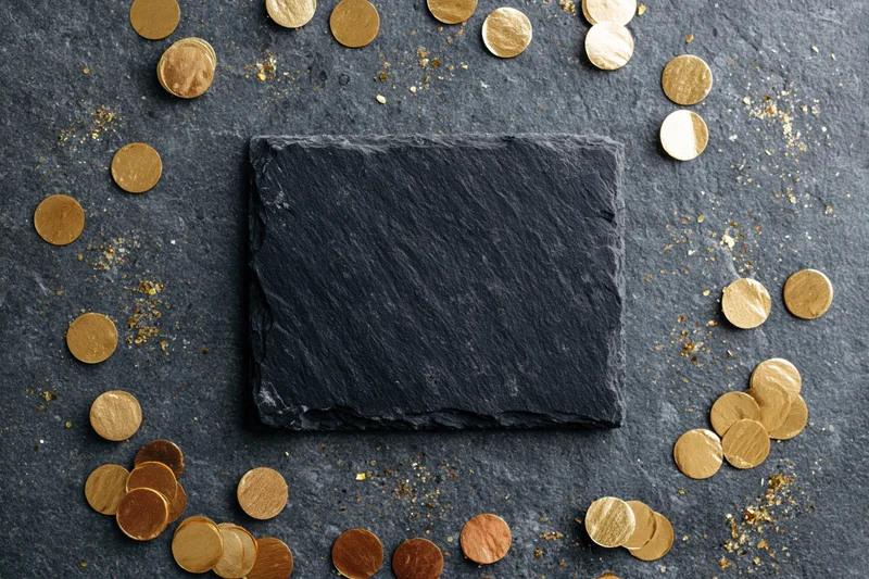Dark slate rectangle centered on textured surface surrounded by scattered gold coins and glitter