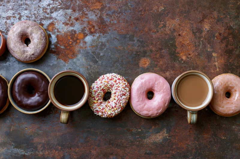 Alternating coffee mugs and glazed donuts arranged in a row on a rusted metal surface