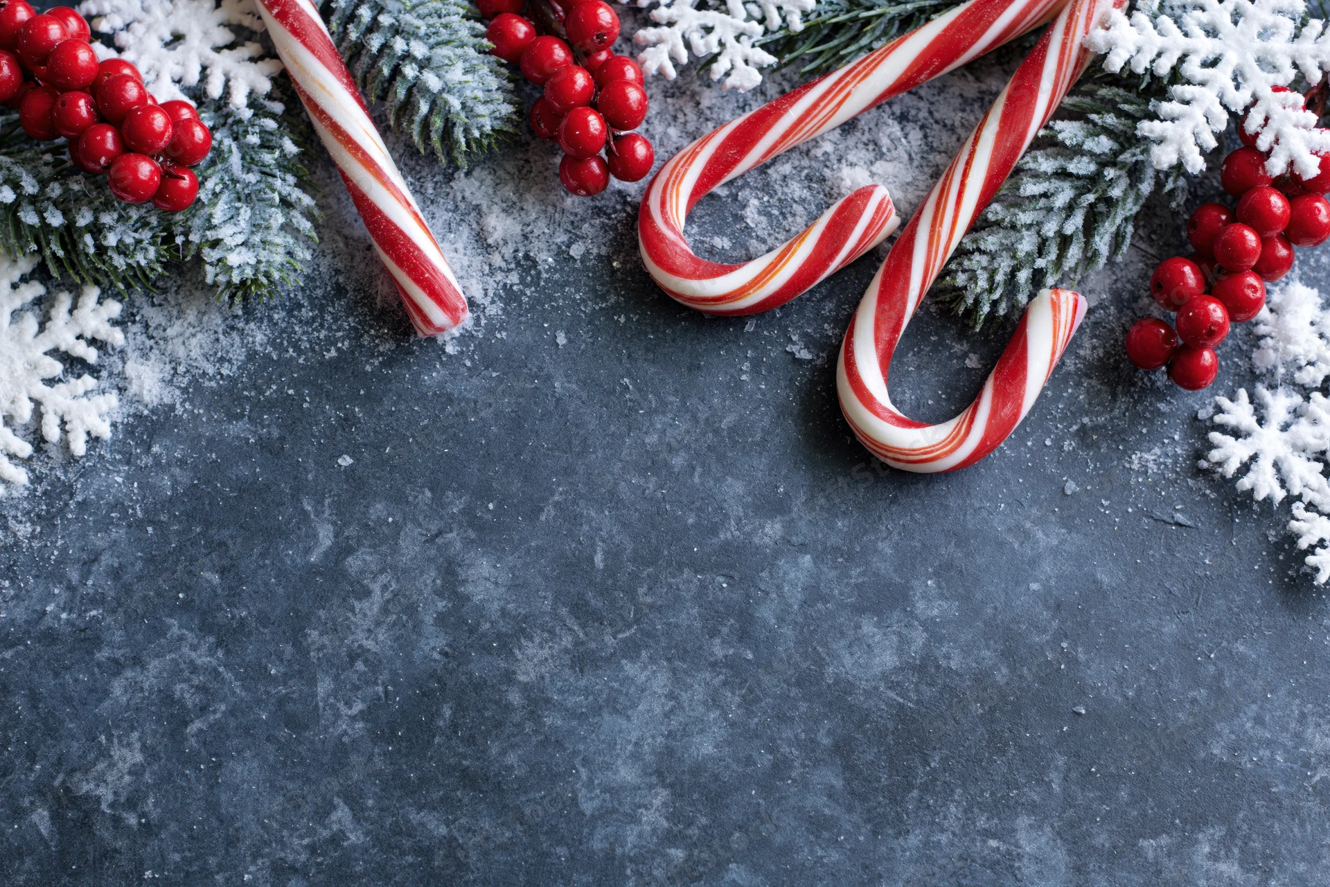 Candy canes and pine branches with red berries and snowflakes arranged along the top edge of a dark textured surface, leaving copyspace below