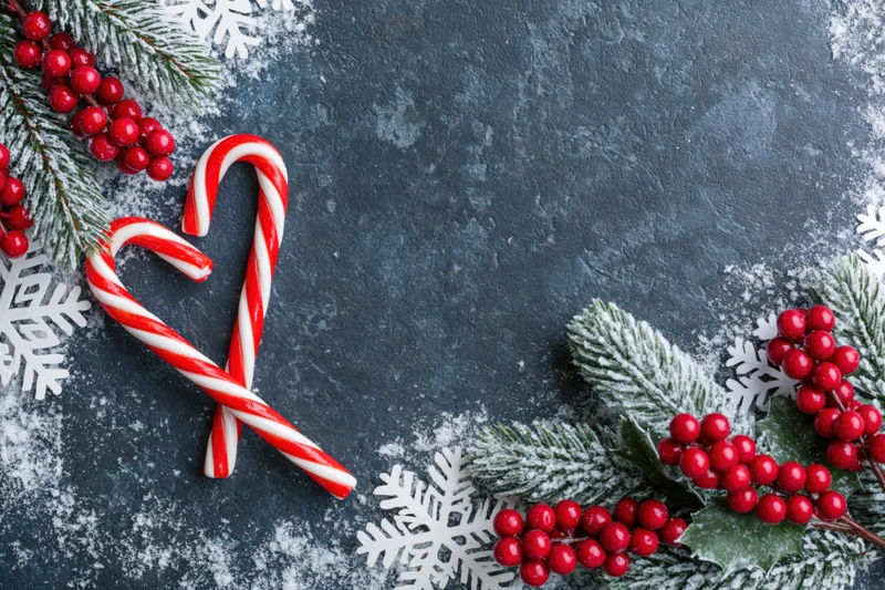 Two candy canes arranged in heart shape with frosted pine branches and red berries on dark textured surface
