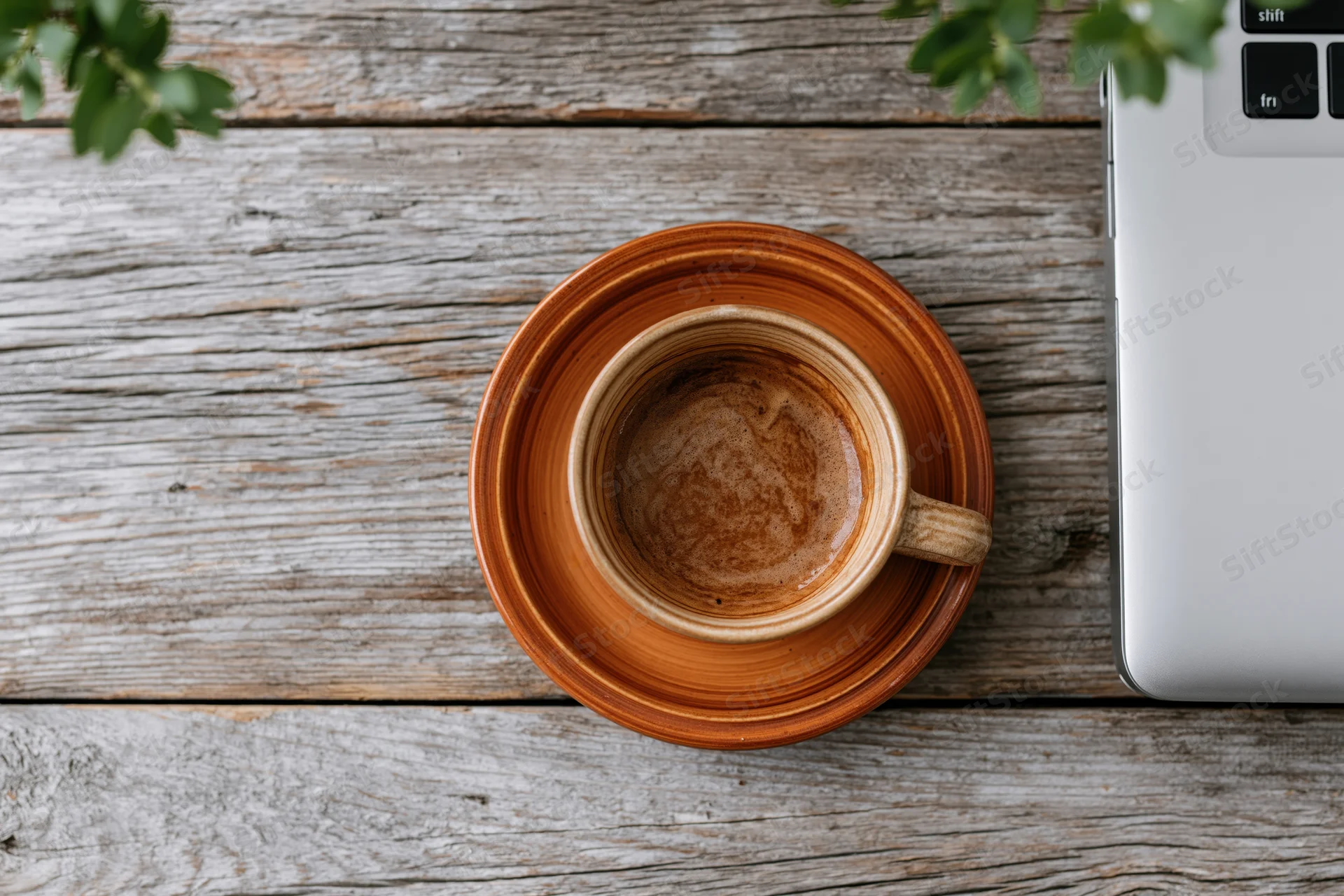 Ceramic coffee cup on wooden table beside laptop with green plant overhead view