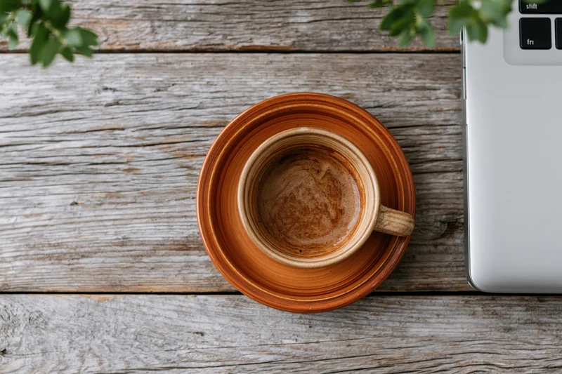 Ceramic coffee cup on wooden table beside laptop with green plant overhead view
