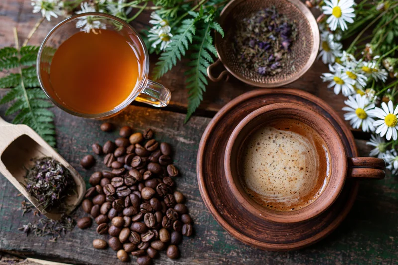 Terracotta coffee cup with foam and scattered beans on wooden table surrounded by flowers and herbs